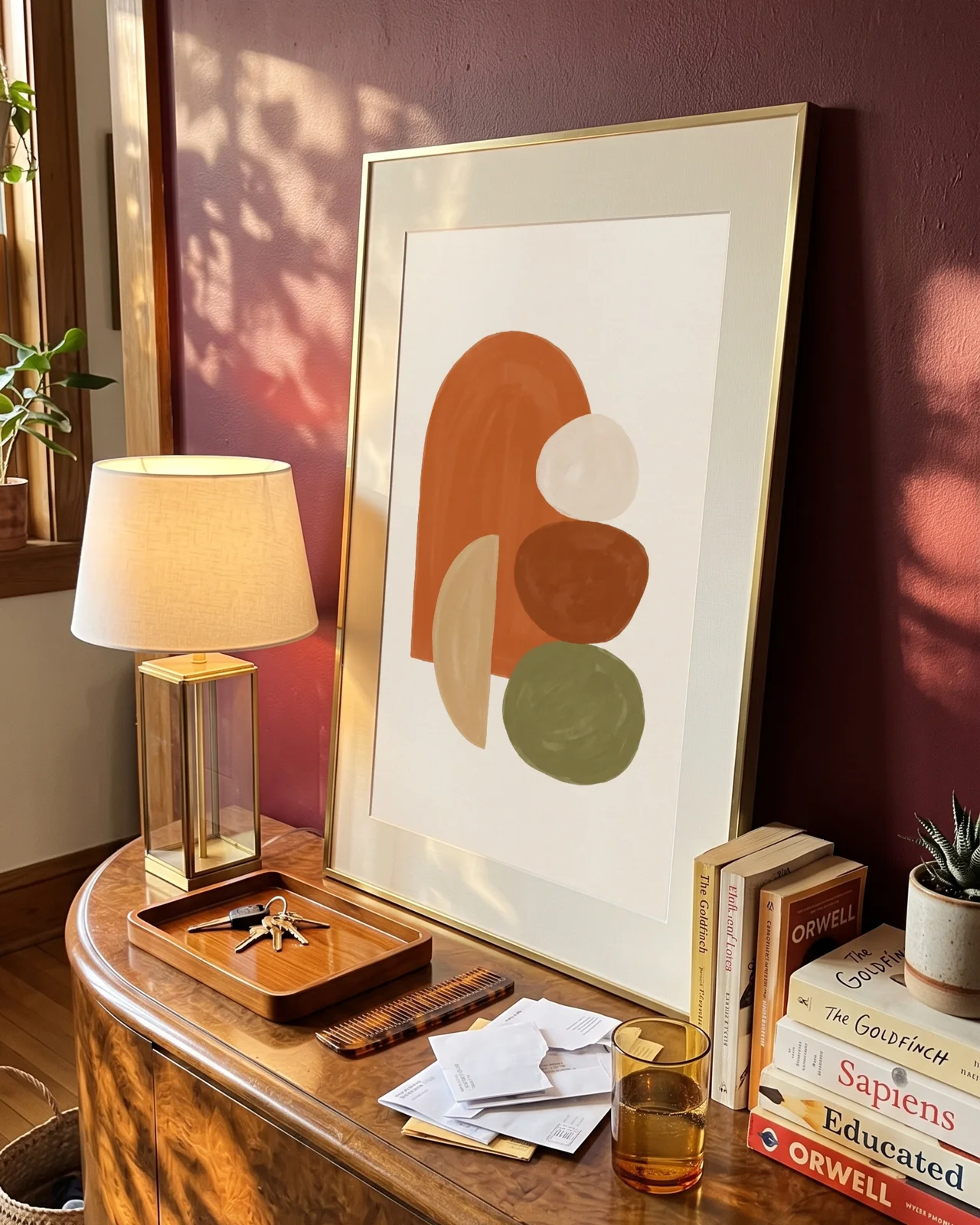 An abstract print of an arch and stacked pebble shapes in a gold frame, leaning against a burgundy wall on a walnut sideboard in window light
